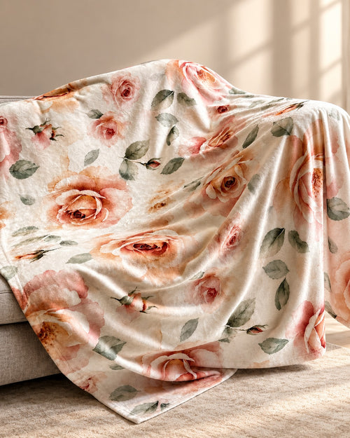 The Bloom with Hope velveteen blanket draped over a cream sofa in warm window light, showing the blush and peach watercolor roses and sage leaves